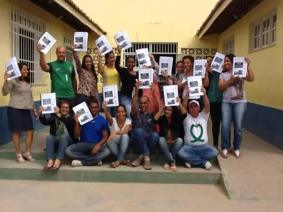 Equipe da Escola Professora Ivani Oliveira reunida em frente à escola, segurando páginas de uma das primeiras produções de material